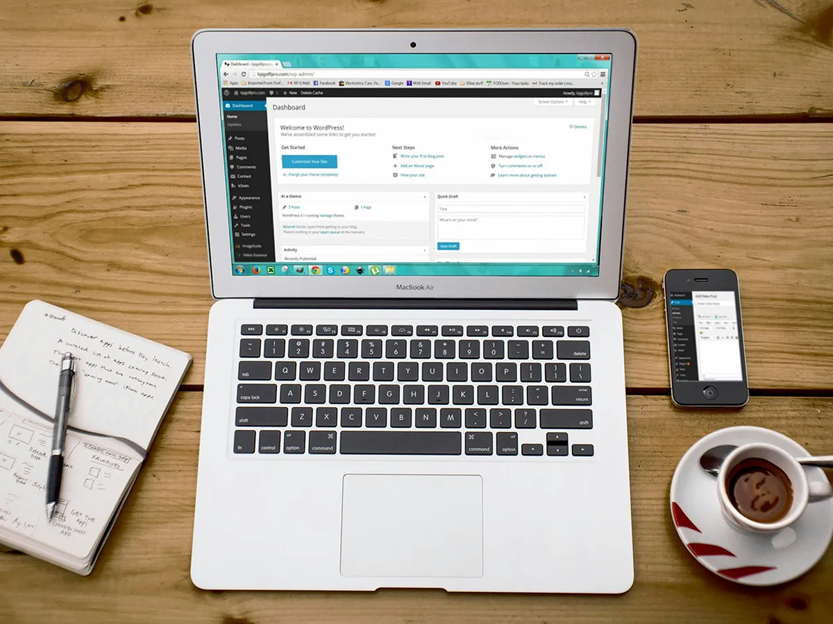 Laptop and smartphone displaying WordPress dashboard with a notebook and coffee on a wooden desk.