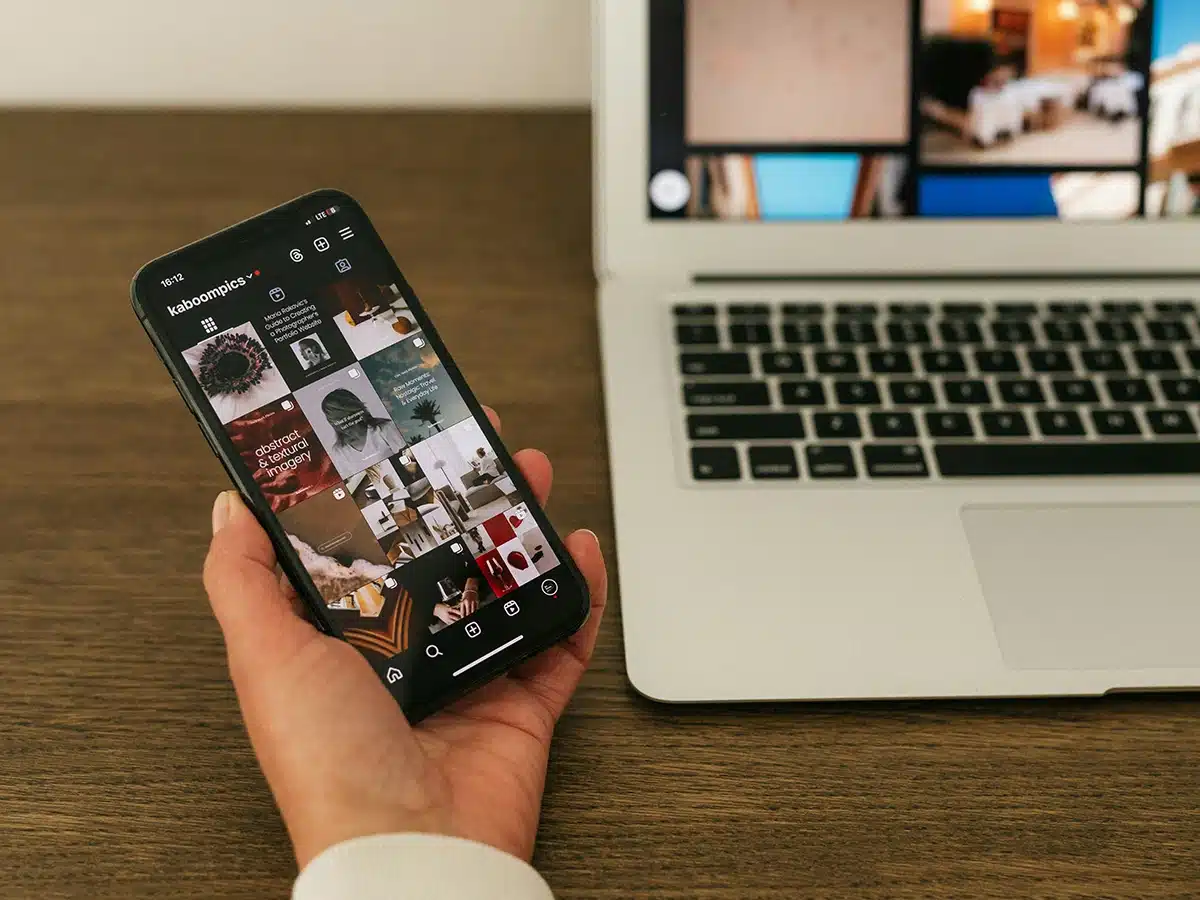 Hand holding a smartphone to check social media trends on Instagram, with a laptop on the right side.
